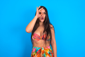 beautiful brunette woman wearing swimwear over blue background doing ok gesture shocked with surprised face, eye looking through fingers. Unbelieving expression.