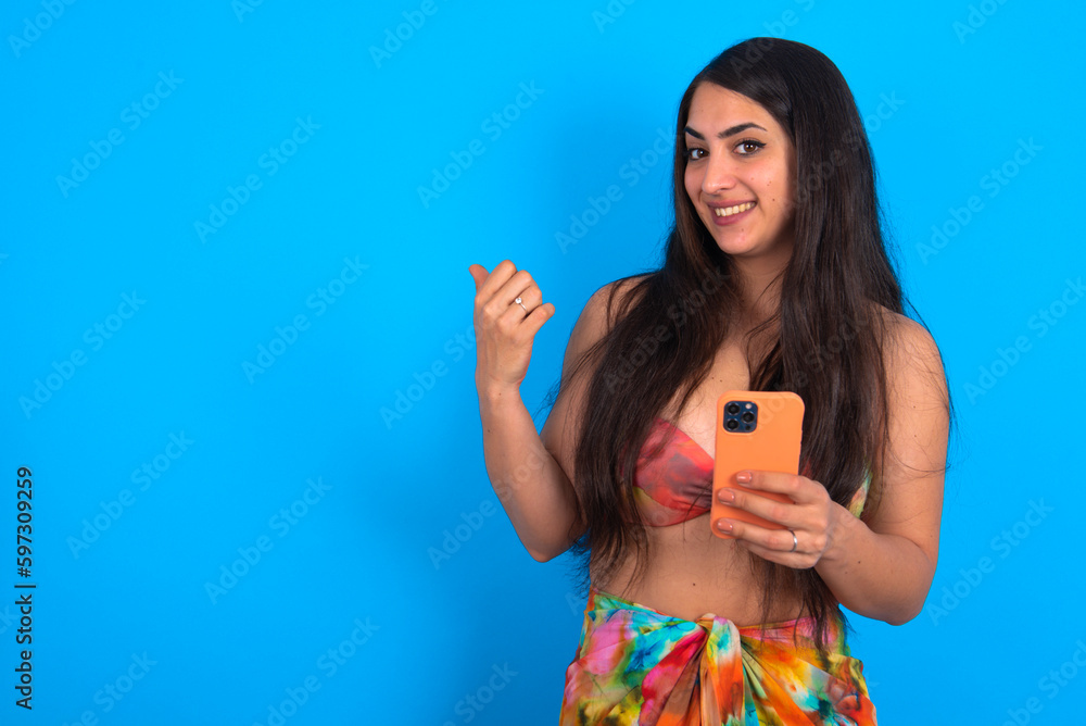 Poster beautiful brunette woman wearing swimwear over blue background using and texting with smartphone poi