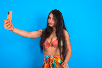 beautiful brunette woman wearing swimwear over blue background smiling and taking a selfie ready to...