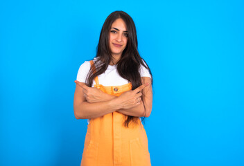 brunette woman wearing orange overalls over blue studio background crosses arms and points at different sides hesitates between two items or variants. Needs help with decision