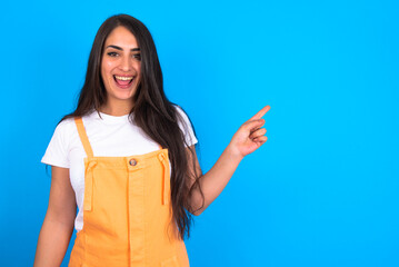 brunette woman wearing orange overalls over blue studio background points aside on copy blank space. People promotion and advertising concept