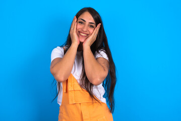Happy brunette woman wearing orange overalls over blue studio background touches both cheeks gently, has tender smile, shows white teeth, gazes positively straightly at camera,