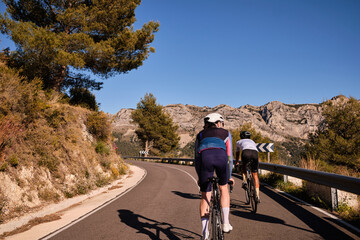 Two cyclists in full cycling gear and helmets ride their road bikes on mountain road at sunset Coll...