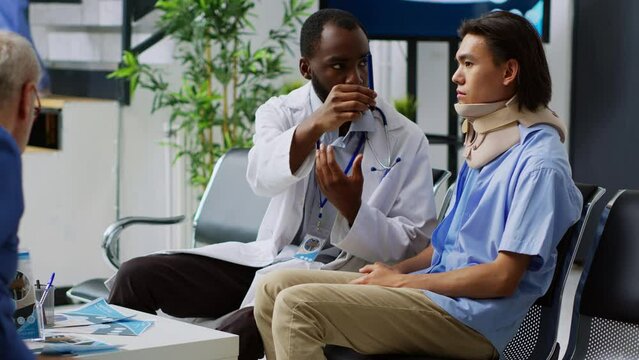 African american medic checking concussion and having medical appointment with asian patient. Person with neck fracture wearing cervical collar in hospital waiting area. Medicine concept