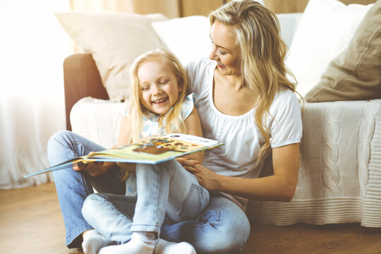 Happy family. Blonde young mother reading a book to her cute daughter while sitting at wooden floor in sunny room. Motherhood concept