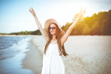 Happy smiling woman in free happiness bliss on ocean beach standing with a hat, sunglasses, and rasing hands. Portrait of a multicultural female model in white summer dress enjoying nature