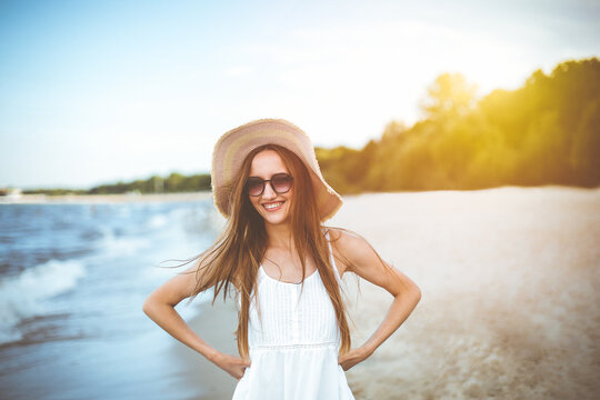 Happy smiling woman in free happiness bliss on ocean beach standing and posing with hat and sunglasses. Portrait of a female model in white summer dress enjoying nature