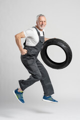 Senior man mechanic in repair shop, holding a new motorcycle tyre, jumping and smiling at camera