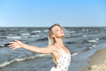 Happy blonde beautiful woman on the ocean beach standing in a white summer dress, raising hands