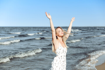 Fototapeta na wymiar Happy blonde beautiful woman on the ocean beach standing in a white summer dress, raising hands