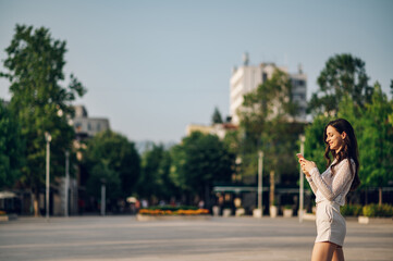 Fototapeta premium A happy young fashionable woman is standing on the city square downtown and writing messages on the phone on a sunny summer day.