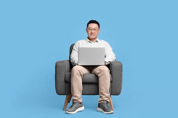 Happy man with laptop sitting in armchair on light blue background