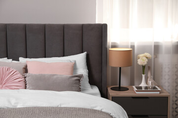 Comfortable bed with cushions, lamp and different decor on bedside table in room. Stylish interior