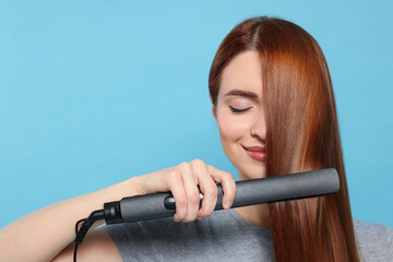 Beautiful woman using hair iron on light blue background, space for text