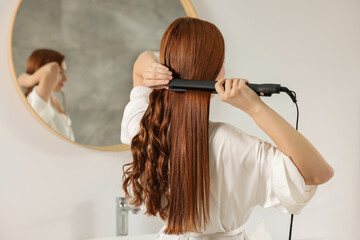 Beautiful woman using hair iron in room, back view. Space for text