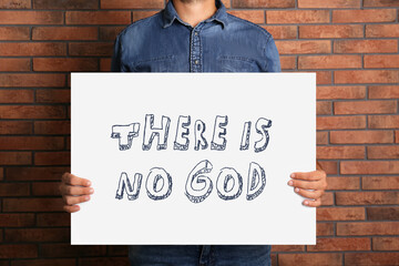 Atheism. Man holding sign with text There Is No God near brick wall, closeup
