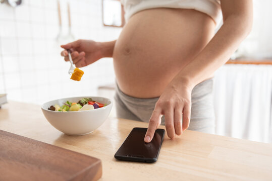 Asian pregnant woman using mobile phone or smartphone while eating healthy salad in kitchen at home.