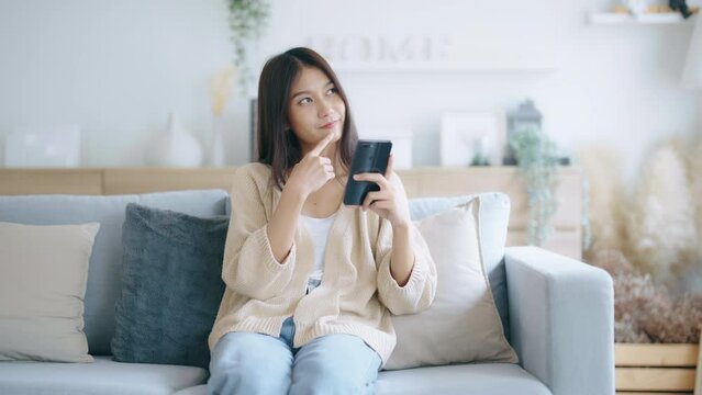 Happy young asian woman relax on comfortable couch at home, typing chat message on smartphone, smiling girl use cell phone chatting, searching information on browser wireless internet, online shopping