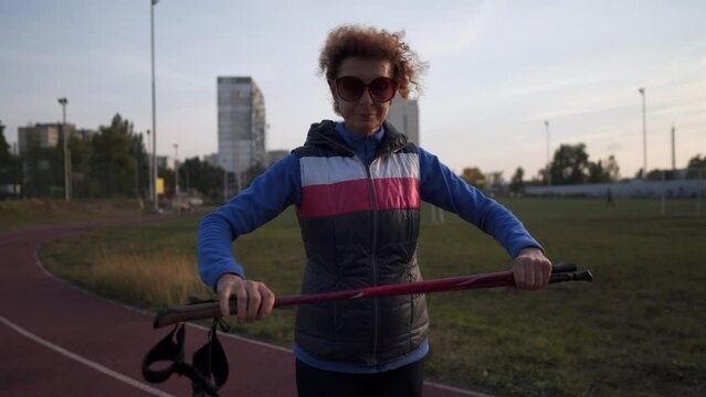 Elderly woman stacks walking sticks at city stadium before workout. Mature athletic female prepares Nordic walking sticks and warms up. Senior athlete gets ready for Nordic walking poles. 