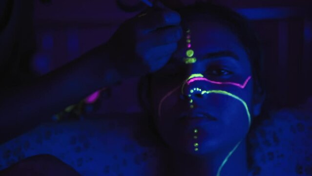 Handheld shot of pretty woman in blue UV light being painted with neon colors in pink and green