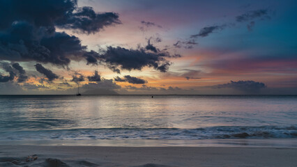 Colorful romantic sunset on a tropical beach. Waves are foaming on the sand. Purple clouds in the sky, illuminated with scarlet, pink, orange. Silhouettes of yachts and islands on the horizon.