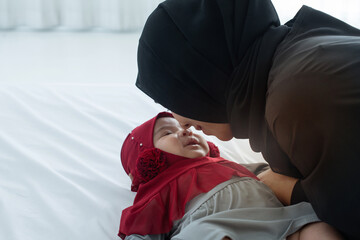 Muslim mom plays and teases happy baby daughter on bed, mother and little baby girl wears hijab, kiss the baby's nose tenderly, view from above, happy loving family concept