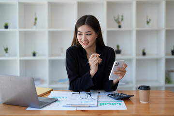 Portrait of a young Asian woman showing a smiling face as she uses her phone, computer and financial documents on her desk in the early morning hours