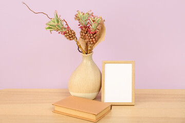 Vase with baby pineapples, book and blank frame on table near lilac wall
