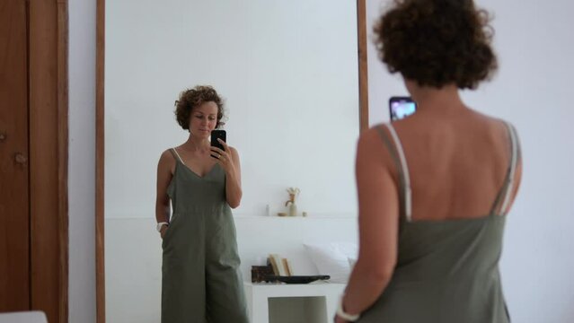 Caucasian woman in front of mirror with phone takes photos of herself, her body, body shape and outfit. Selfie in front of mirror, an attractive woman takes photos, selfie on a phone camera, rear view