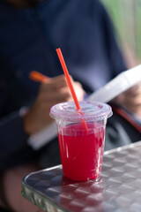 In the foreground, a cold serving of brightly colored fruit punch sits on a patio table. In the background, a person writes in their diary, enjoying a moment out and about during the warm summer.