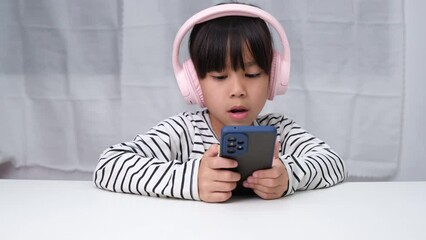 Cute elementary school girl wearing headphones holding a smartphone. Happy Asian girl studying online on smartphone or homeschooling, listening to music or playing games.