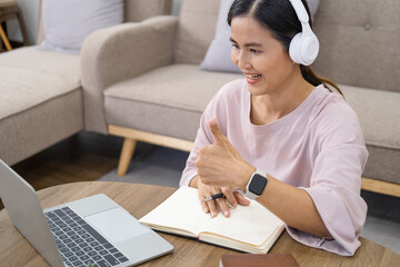 Asian woman teacher in headphones show sight thumbs up, is using a laptop and communicates on the Internet at home. homeschooling, distant learning, online e