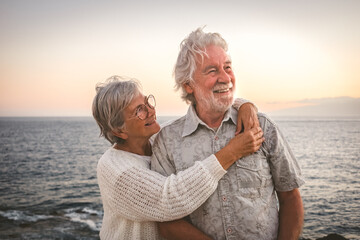 Cheerful romantic senior couple embraced at the sea at sunset light - old people outdoors enjoying...