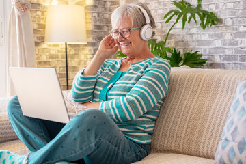 Beautiful caucasian senior woman wearing headphones sitting on sofa at home looking video on laptop - concept of relaxed lady enjoying retirement and technology