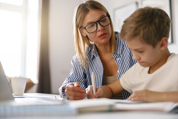 Fototapeta premium Schoolboy sitting at table and writing in copybook on online lesson while his mom helping him