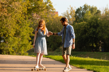 Obraz premium Young man embracing and helping girlfriend riding longboard on asphalt walkway between trees