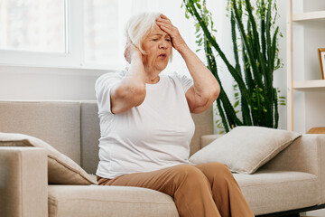 Elderly woman severe pain in the head sitting on the couch, health problems in old age, poor quality of life. Grandmother with gray hair holding her head, migraine and high blood pressure