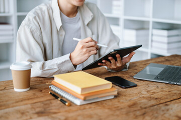 Young Asian man student working on his project assignment, using laptop computer and tablet to search an online informations..