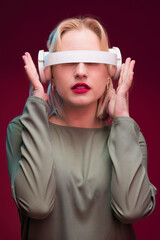 A goofy blond woman playing around with headphones and looking trough it while standing isolated on magenta background. Viva magenta, color of the year.
