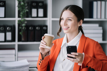 Businesswoman sitting in modern office using smartphone playing social media and enjoying coffee