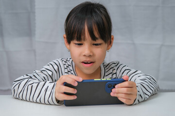 Cute little girl using smartphone and smiling while sitting at table in house. Happy Asian girl holding phone in hands, online learning or homeschooling, listening to music or playing games.