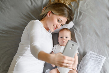 Happy cute mother and baby in bed, looking at mobile phone camera, taking selfie. Young family communication online, video call, technology, communication concept.