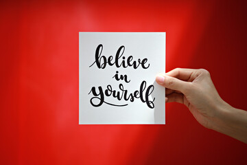 Hand holding a message card with positive phrase "Believe in Yourself" on a blur red background