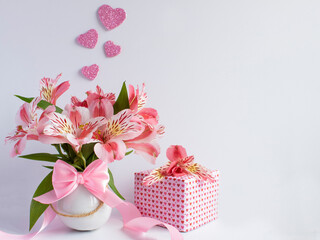 bouquet of pink flowers in a small white vase and a gift next to it with paper with small pink hearts and a flower on top,on the flowers you can see four four pink glitter hearts on a white background