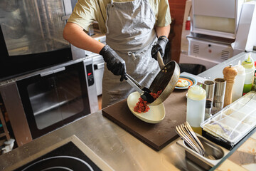 Chef putting ready dish from frying pan into the plate before serving to client in restaurant