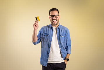 Handsome male customer with hand in pocket showing credit card and laughing ecstatically on isolated beige background. Cheerful young man recommending online shopping and cashless payment - obrazy, fototapety, plakaty