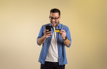 Male customer with credit card and smart phone screaming excitedly after winning rewards for online shopping. Cheerful young man buying through mobile apps on beige background