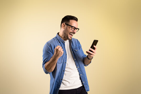 Male entrepreneur screaming ecstatically and pumping fist while reading good news over smart phone. Cheerful young man celebrating achievement while standing on beige background