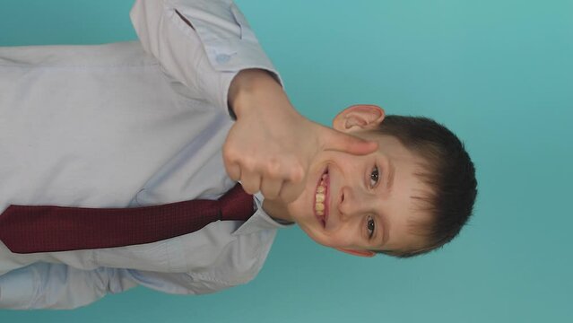 handsome european schoolboy in a shirt and tie looks at the camera and shows dest thumb up, blue background, studio shot.vertical video. sign of consent and approval
