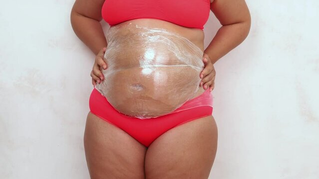 Female hands pound on wrapping with cling film stomach. Overweight woman in red underwear on white background. Anti cellulite fat burning body wrap, therapy. Skincare, weightloss. Plus size people.